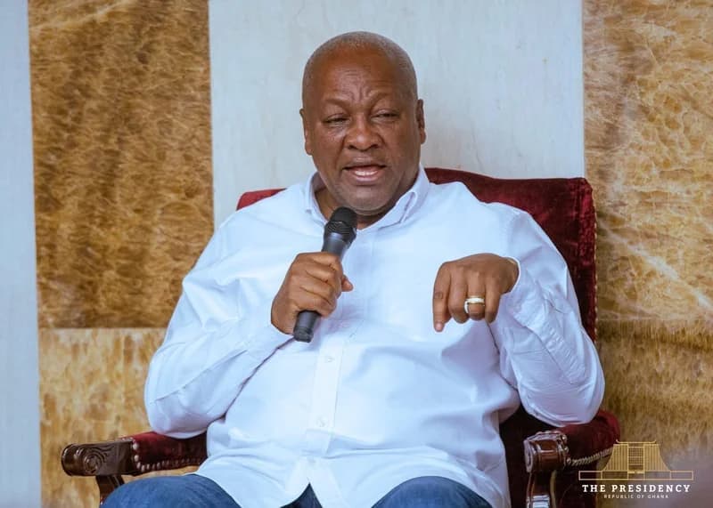 President John Dramani Mahama is captured speaking into a microphone while seated in a formal indoor setting. He is dressed in a white long-sleeved shirt and blue trousers, gesturing with his hand as he delivers a statement.