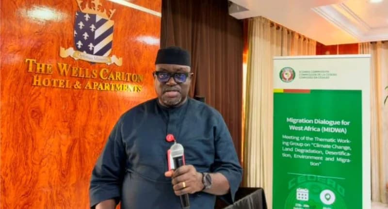 A man in dark traditional attire and glasses speaks into a microphone during an ECOWAS conference. Behind him, a wooden wall displays the logo for The Wells Carlton Hotel & Apartments, while a green banner to his right details a 'Migration Dialogue for West Africa (MIDWA)' meeting focused on climate change and land degradation.