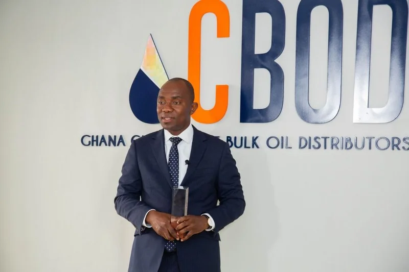 A man in a navy blue suit and patterned tie stands in front of the Ghana Chamber of Bulk Oil Distributors (CBOD) logo while holding a small rectangular award. He appears to be speaking or posing during an official function related to the energy sector.