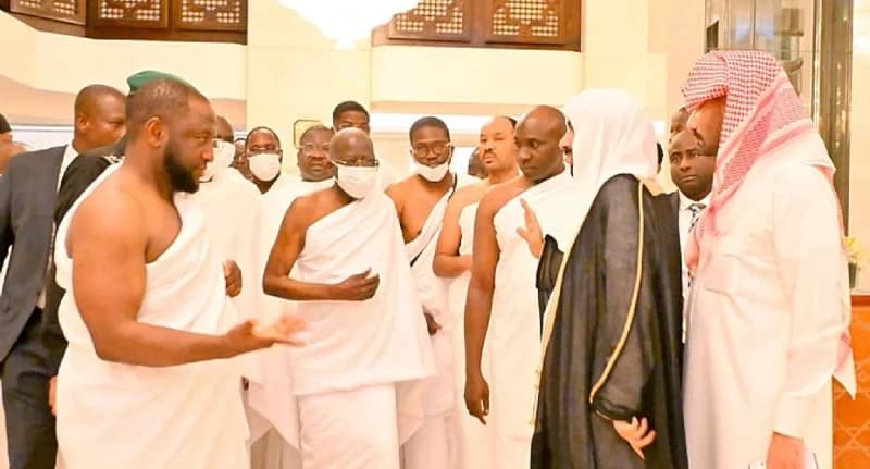 Nigerian President Bola Tinubu, wearing a white Ihram and a face mask, is pictured during his Umrah pilgrimage in Saudi Arabia surrounded by his entourage and Saudi officials. The group is standing in a brightly lit indoor space, appearing to engage in conversation while dressed in traditional religious attire for the pilgrimage.