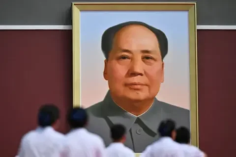 AFP via Getty Images Large framed portrait of Mao Zedong hanging on a red wall, viewed from below with several people in light-coloured clothing standing and facing the portrait in the foreground.