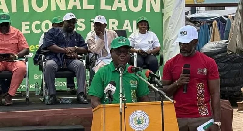 A man in a green polo shirt and matching cap addresses a crowd from a wooden podium bearing the seal of Ghana's Ministry of Foreign Affairs. Several officials are seated behind him on a stage in front of a green backdrop promoting an ECOWAS initiative.